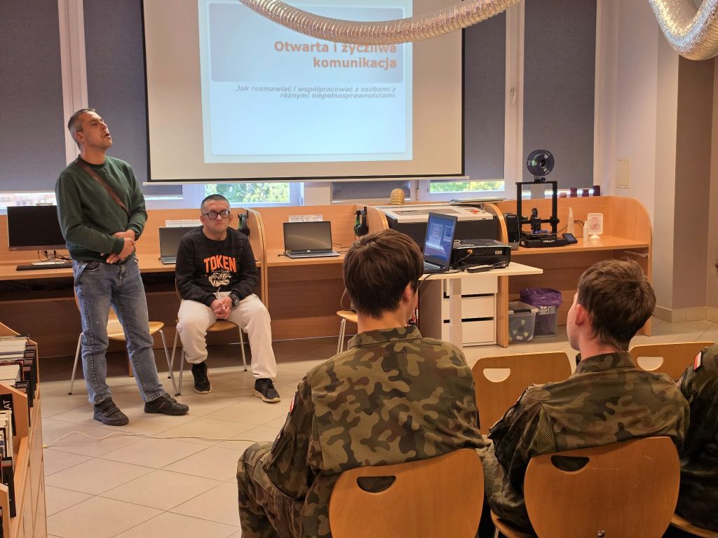 Dwóch prowadzących znajduje się przed grupą uczniów w mundurach. W tle widoczny jest ekran z prezentacją dotyczącą komunikacji.