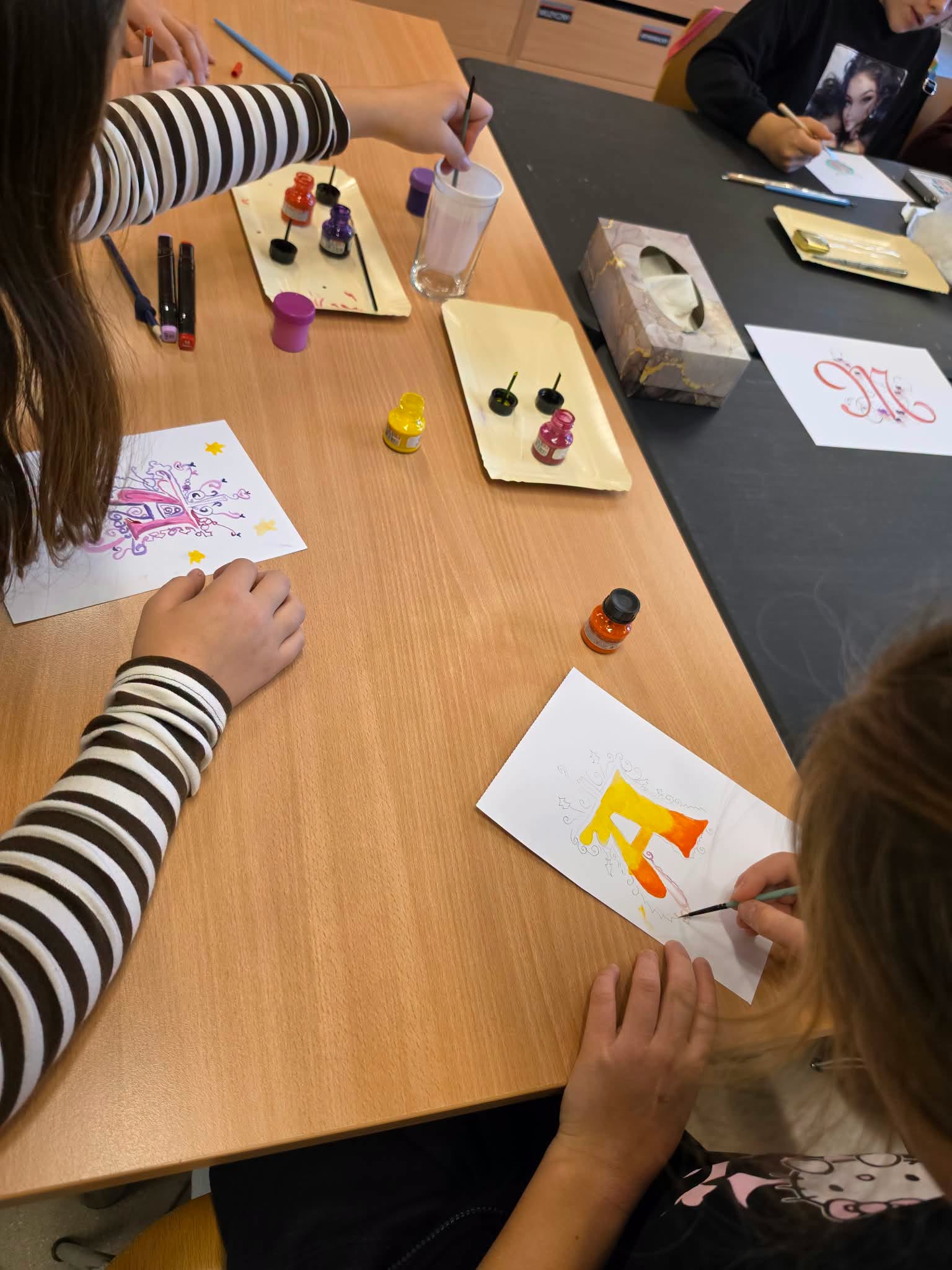Uczestniczki warsztatów kaligrafii ozdabiają dekoracyjne inicjały kolorowymi tuszami i farbami. Na stole leżą pędzelki, przybory plastyczne oraz kartki z gotowymi i powstającymi pracami.