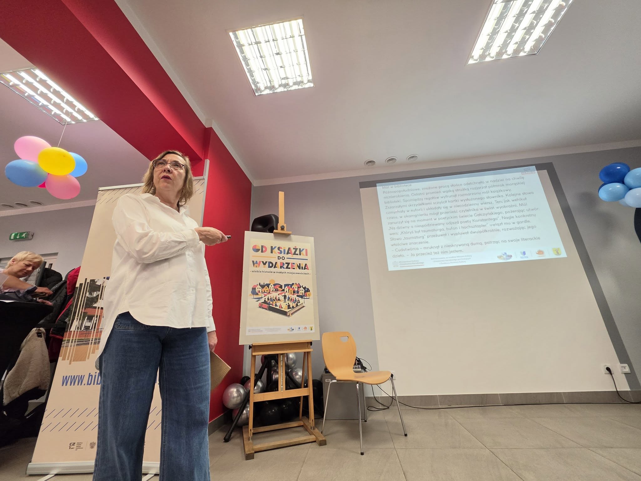 Kobieta prowadzi Dyktando Międzypokoleniowe w Miejskiej Bibliotece Publicznej w Morągu. Stoi przed ekranem, na którym wyświetlony jest tekst „Mól w bibliotece”. Obok znajduje się plakat projektu „Od książki do wydarzenia”.