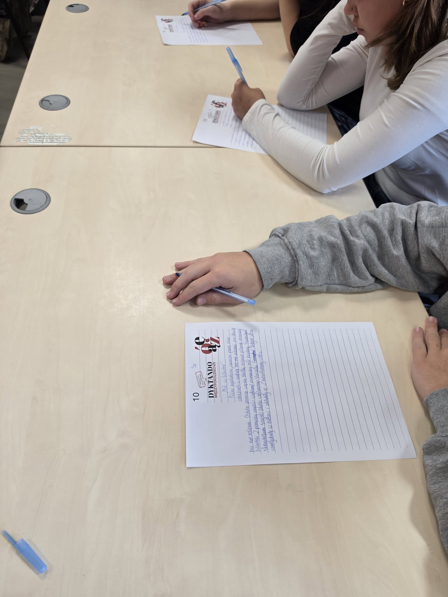 Zbliżenie na arkusze Dyktanda Międzypokoleniowego wypełniane przez młodych uczestników. Na pierwszym planie leży częściowo zapisany tekst, a przy sąsiednim miejscu uczennica pisze niebieskim długopisem.