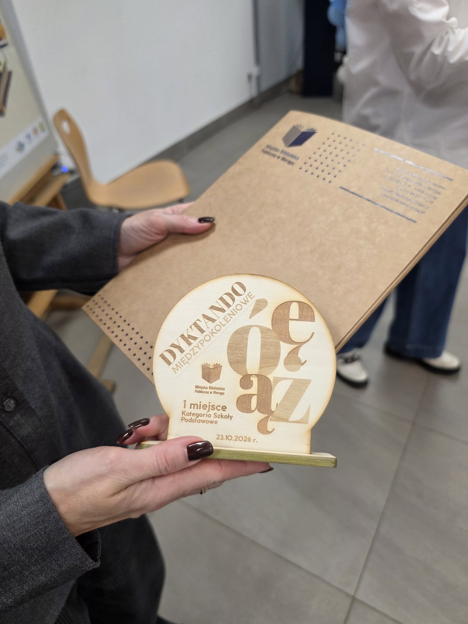 Zbliżenie na drewnianą statuetkę przygotowaną na ploterze laserowym w Pracowni Orange. Nagroda za pierwsze miejsce w kategorii szkół podstawowych ma wygrawerowany tytuł „Dyktando Międzypokoleniowe”, ozdobne litery z polskimi znakami, logo biblioteki i datę wydarzenia. W tle widoczna jest pamiątkowa tabliczka.
