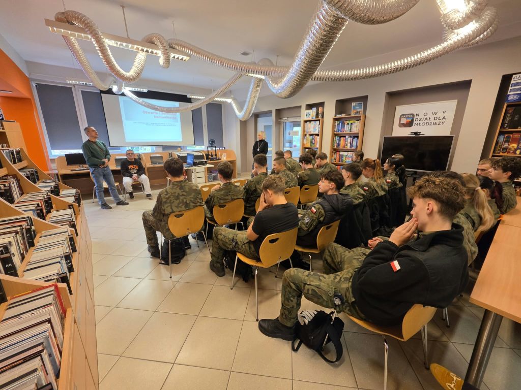 Dwóch prowadzących znajduje się przed grupą uczniów w mundurach. W tle widoczny jest ekran z prezentacją dotyczącą komunikacji.