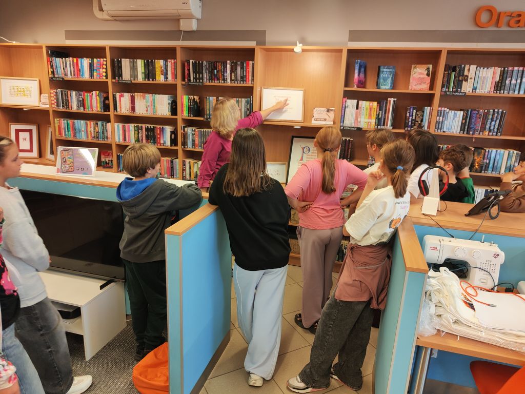 Uczestnicy warsztatów stoją przy regale z książkami, a prowadząca wskazuje oprawioną pracę.