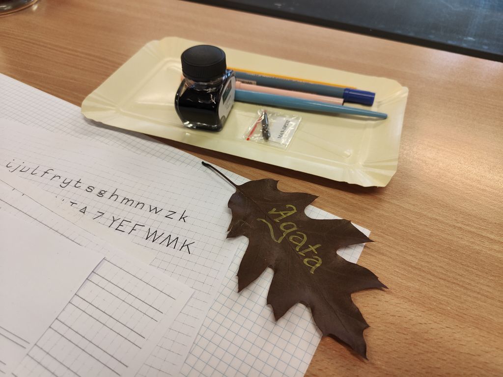 Kałamarz, pióra i kartka do ćwiczeń kaligrafii obok liścia z imieniem „Agata”.