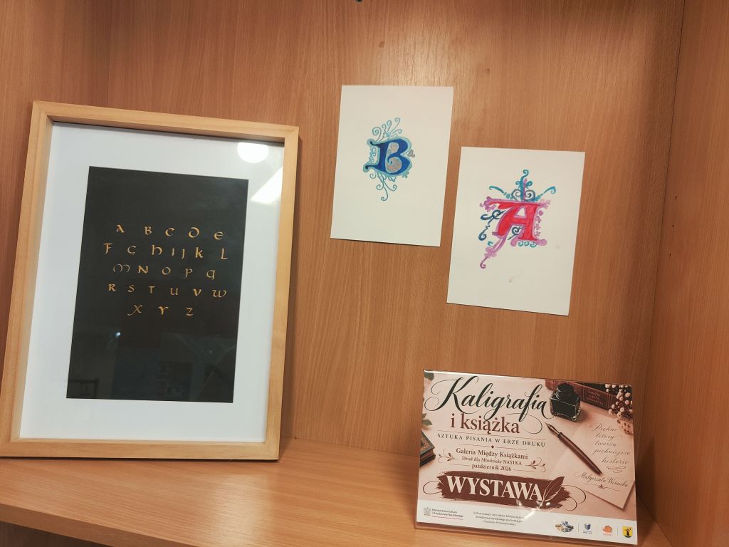 Drewniana półka z wystawą kaligrafii. W ramce znajduje się alfabet zapisany ozdobnym pismem, obok dwie kolorowe prace z inicjałami „B” i „A” oraz plansza informująca o wystawie.