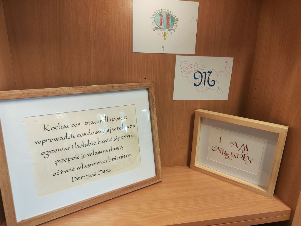 Fragment wystawy kaligrafii na drewnianej półce. W dwóch ramkach znajdują się kaligraficzne napisy, a nad nimi dwie kolorowe prace z ozdobnymi inicjałami.