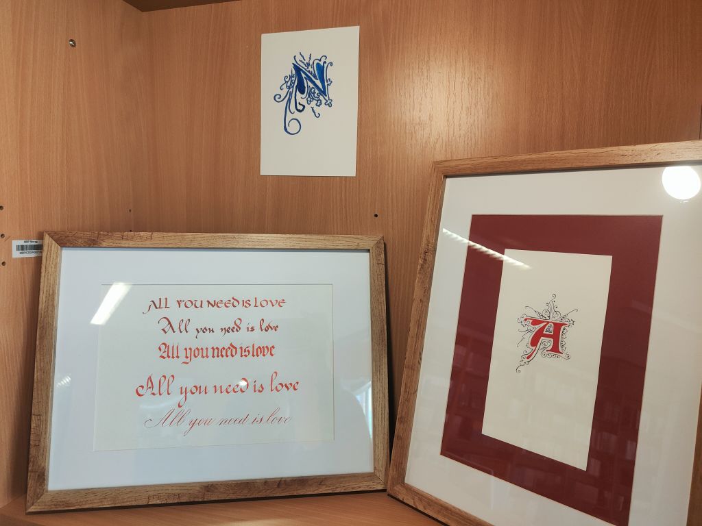 Drewniana półka z trzema pracami kaligraficznymi. W ramce po lewej widoczny jest napis „All you need is love” zapisany różnymi stylami, po prawej ozdobny inicjał „A”, a nad nimi niebieski inicjał „N”.