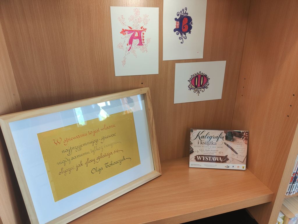 Fragment wystawy kaligrafii z pracą w ramce zawierającą cytat Olgi Tokarczuk oraz trzema kolorowymi, ozdobnymi inicjałami zawieszonymi nad nią. Obok stoi plansza informacyjna wystawy.