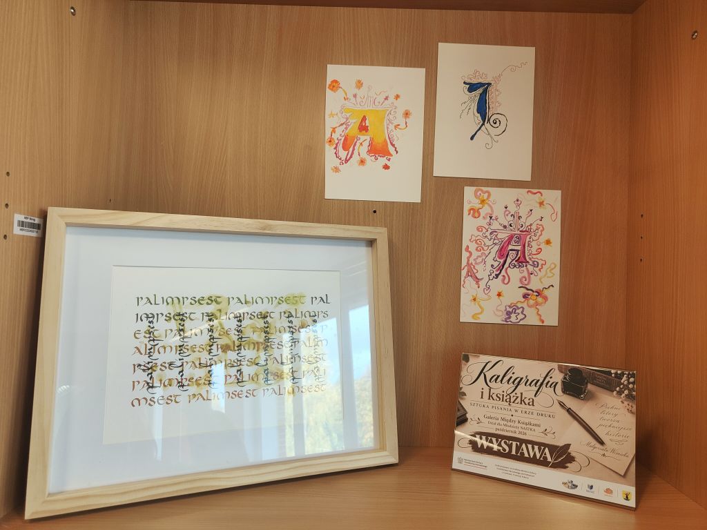 Drewniana półka z oprawioną pracą kaligraficzną oraz trzema kolorowymi inicjałami wykonanymi na białych kartkach. Po prawej stoi plansza informująca o wystawie.