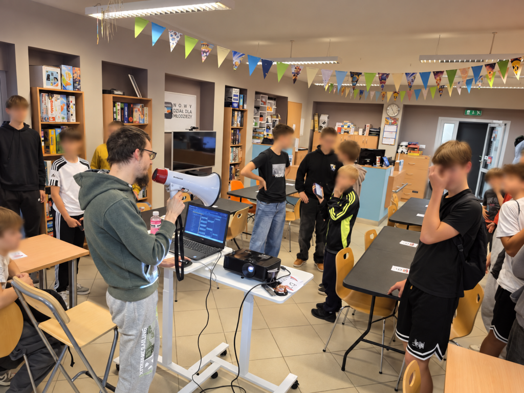 Mężczyzna z megafonem stoi przy laptopie i projektorze, wokół niego grupa uczestników.