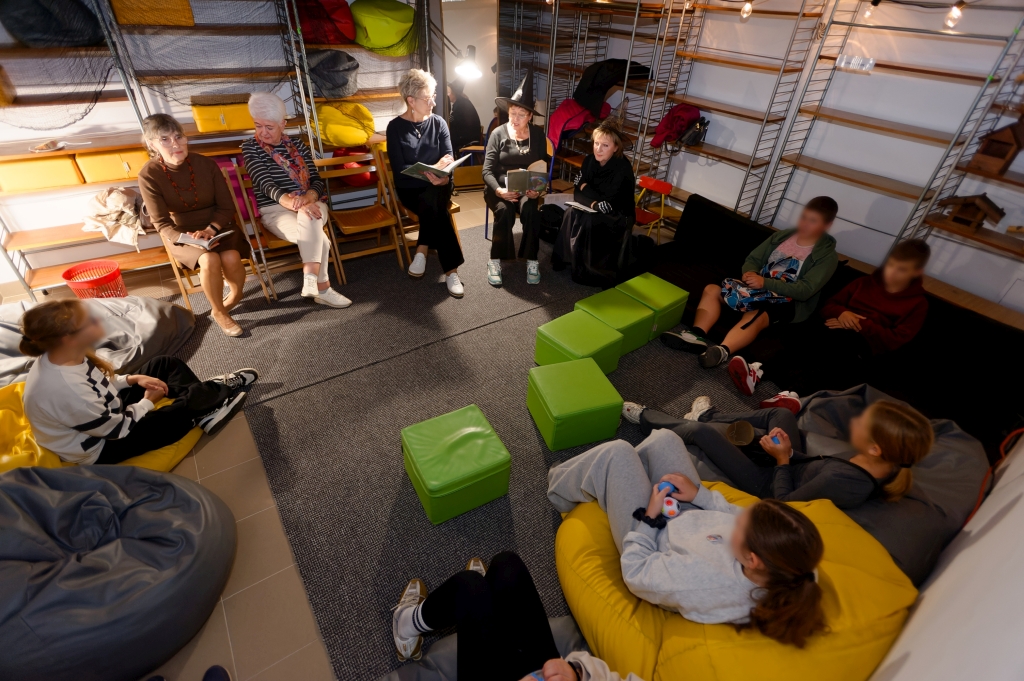 Grupa uczestników słucha utworów literackich czytanych przez kobiety w bibliotecznej piwnicy.