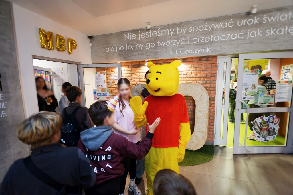 Dzieci witają się z maskotką Kubusia Puchatka w holu biblioteki.