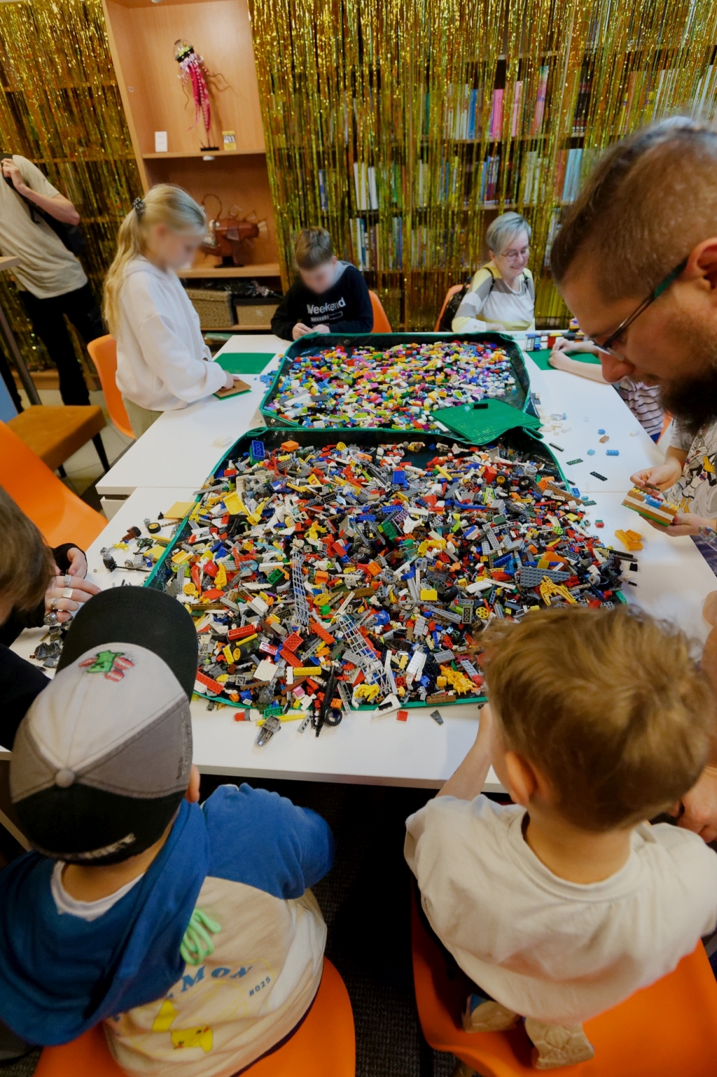 Dzieci i dorośli budują konstrukcje z klocków LEGO przy wspólnym stole.