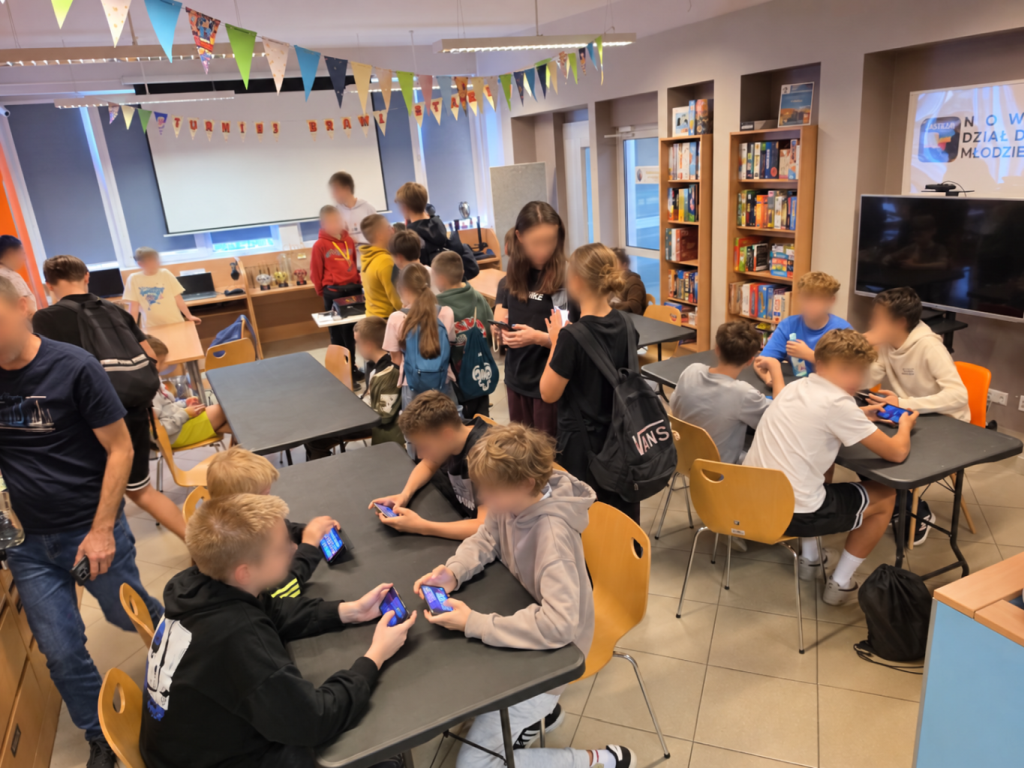 Grupa osób siedzi i stoi przy stołach, korzystając ze smartfonów.