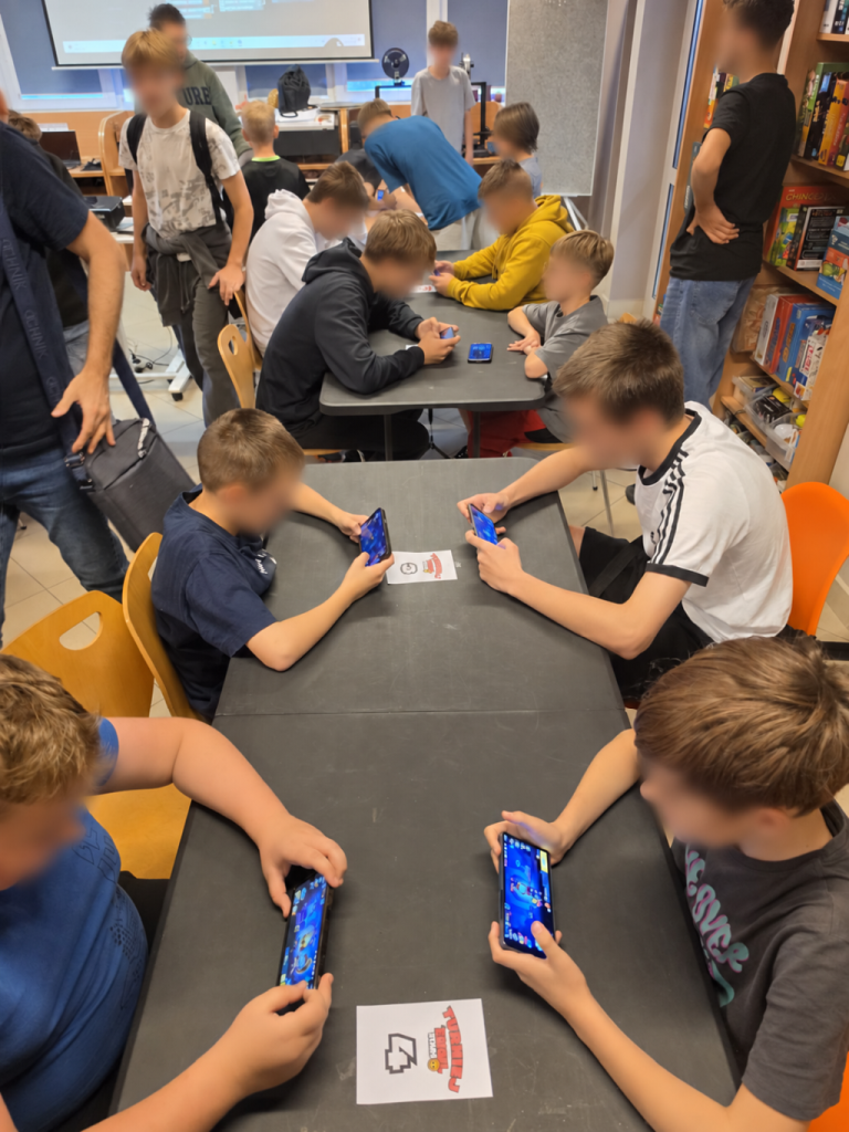Grupa młodzieży siedzi i stoi przy stołach, grając na smartfonach.