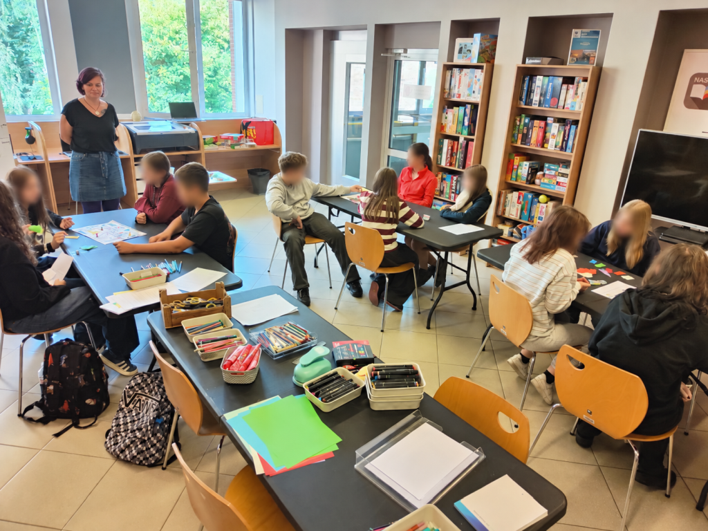 Młodzież pracuje w grupach przy stołach, a prowadząca stoi z przodu sali.
