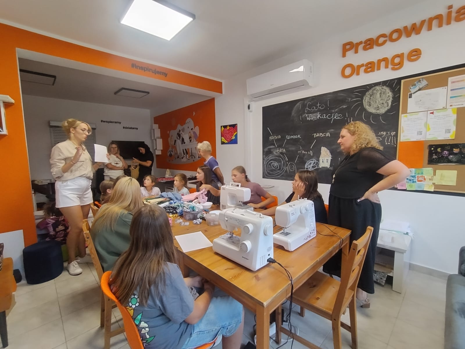 Kobiety przy stole z maszynami do szycia, Pani prowadząca tłumaczy jak uszyć scrunchie