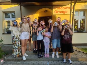 zdjęcie przedstawia grupę uczestników warsztatów, którzy trzymają własnoręcznie wykonaną scrunchie gumkę na twarzy
