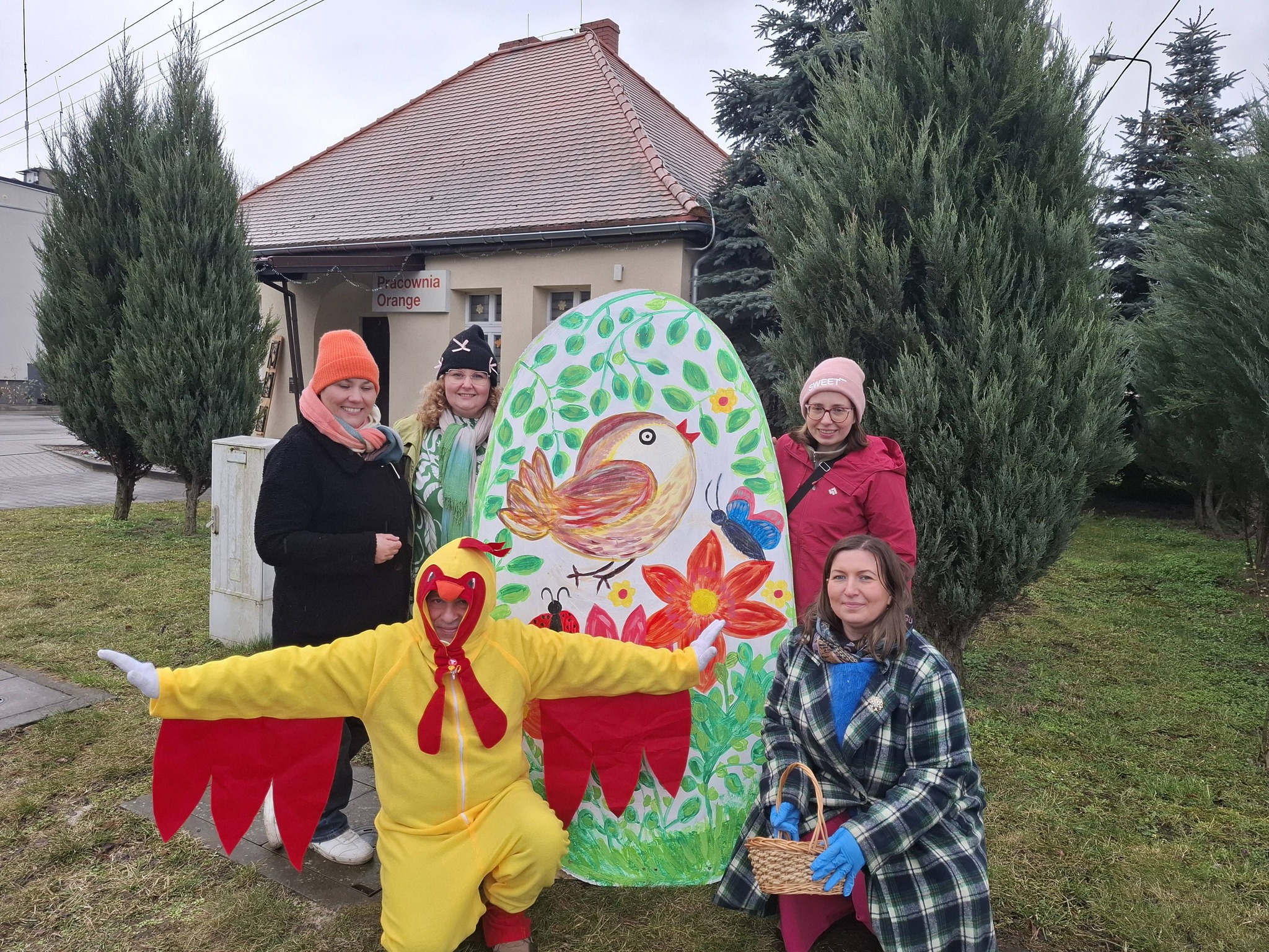 na zdjęciu duży kurczak, łącxznie 5 osób dorosłych, które pozują do zdjęcia na tle dużego jajka z drewna, pomalowanego farbami, na jajku widac ptaszka i liście