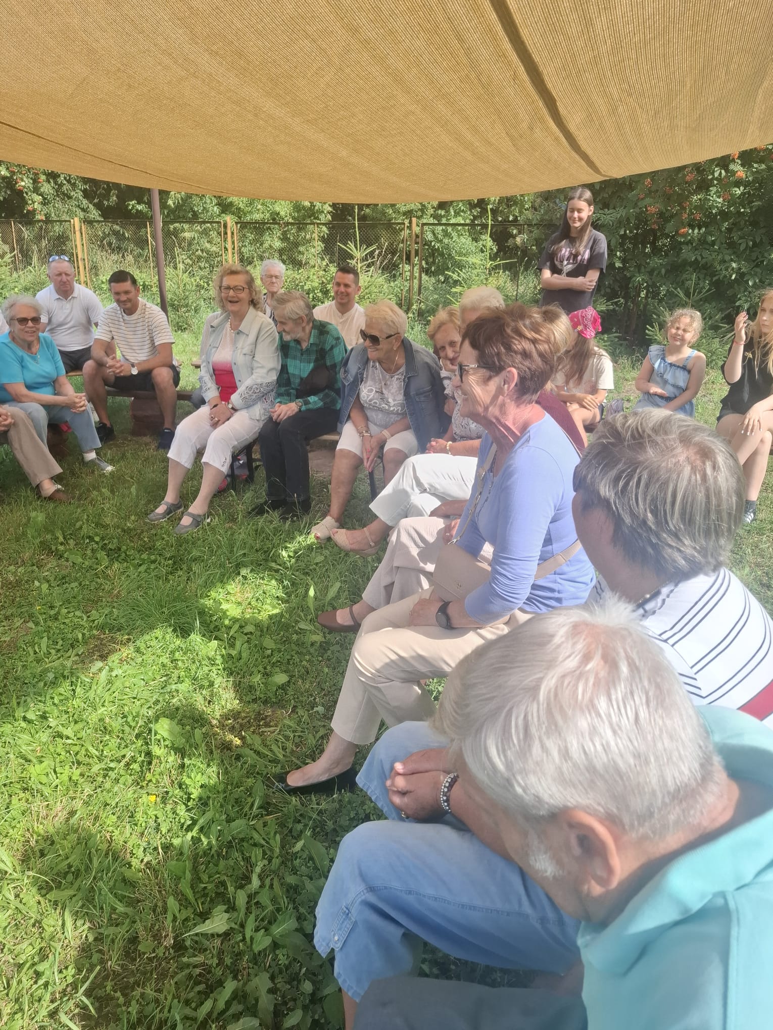 Grupa seniorów siedzących na ławkach pod namiotem.