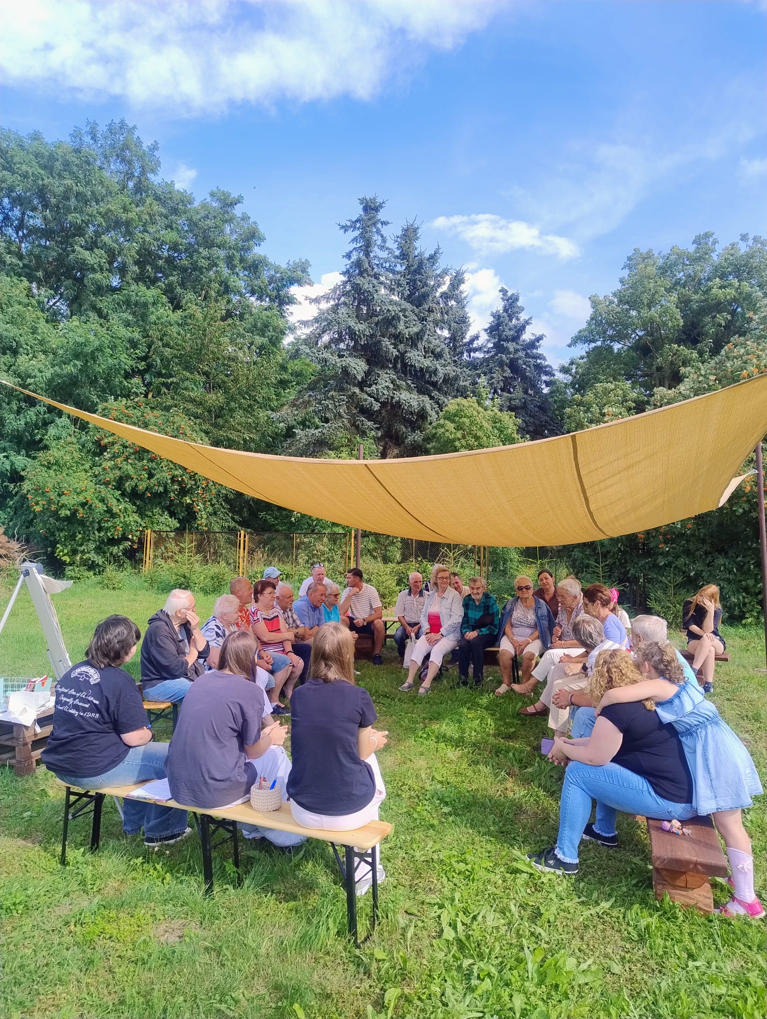 Grupa osób siedzi na ławkach pod namiotem.