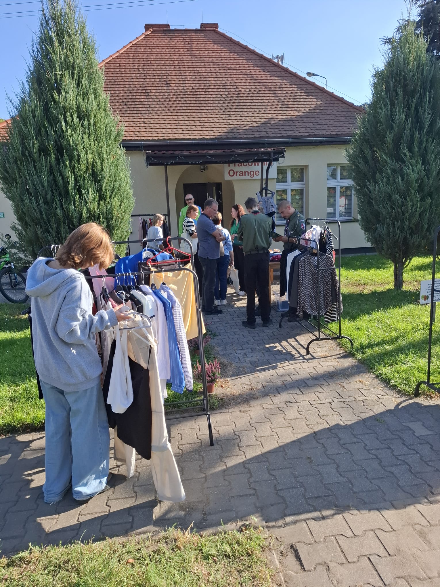 Grupa osób przed Pracownią Orange, wzdłuż chodnika stoją wieszaki z ubraniami.