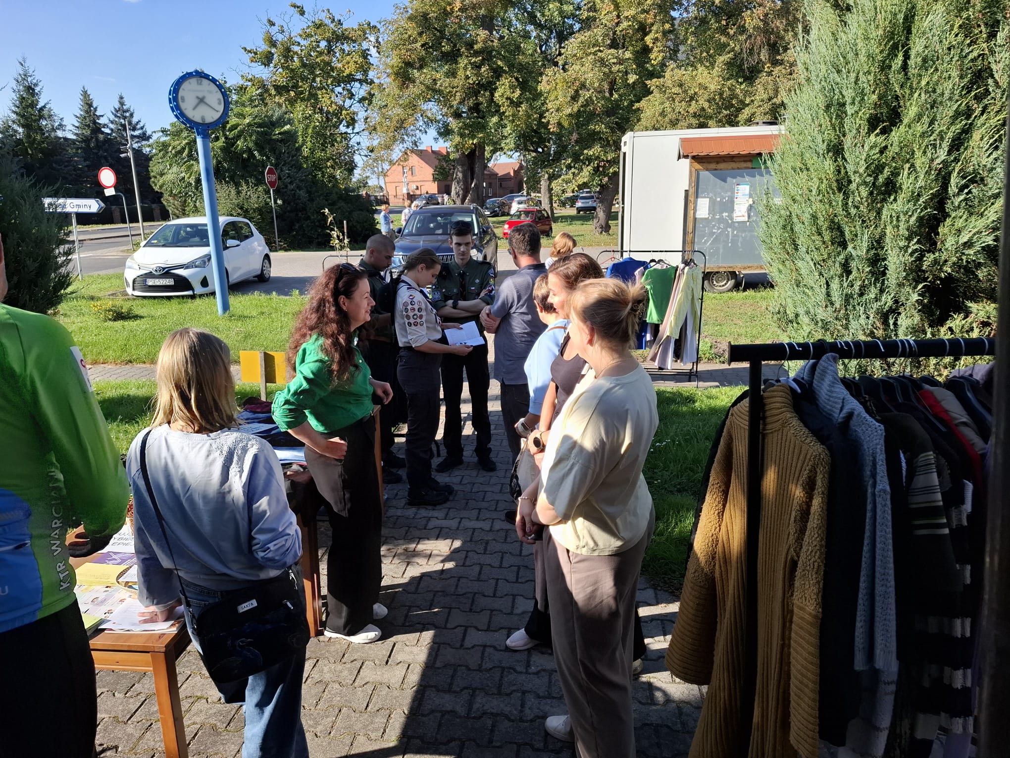 Grupa osób zebranych przy wieszakach z ubraniami, które ustawione są na dworze.