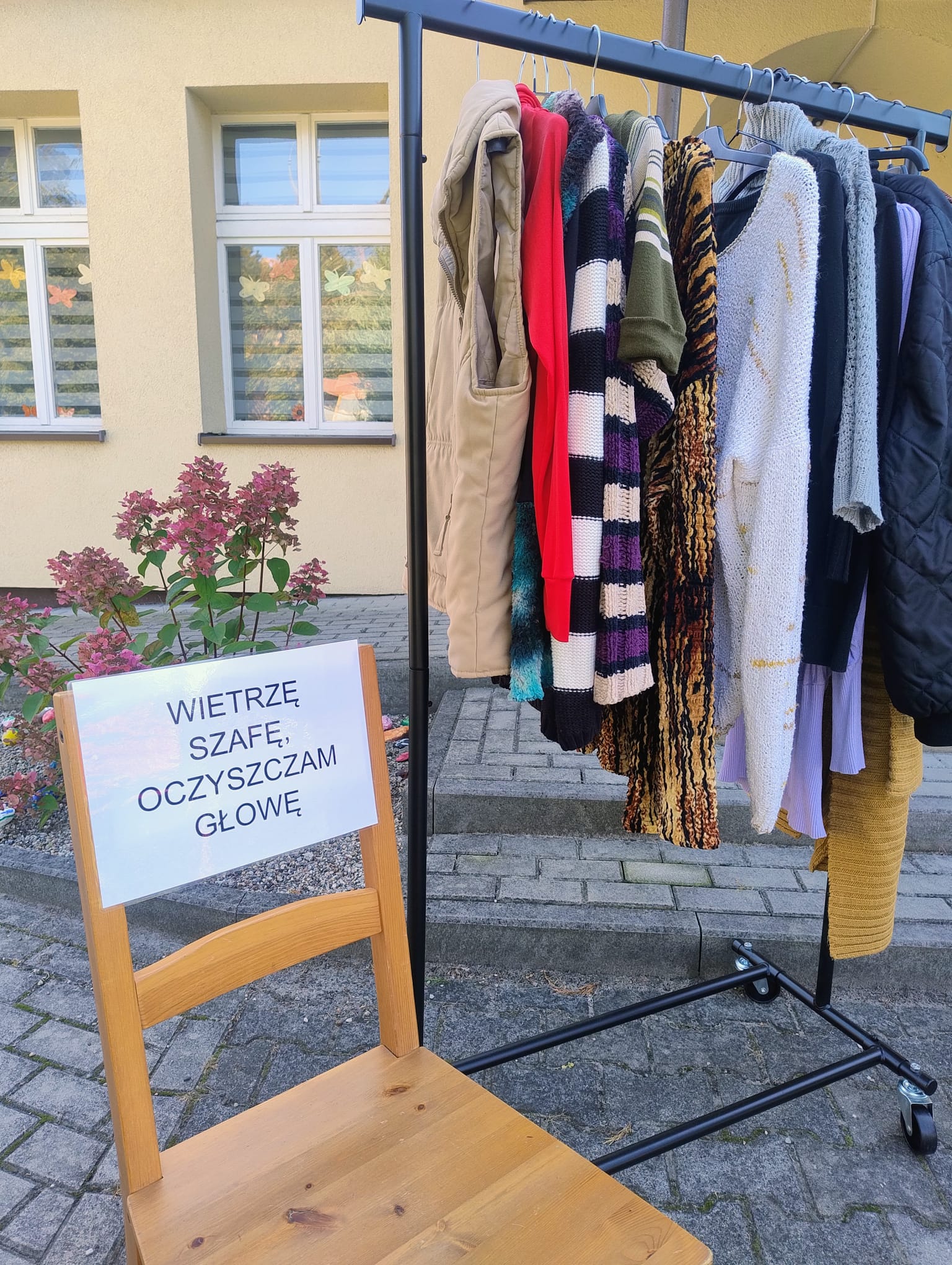 Na zdjęciu w lewym dolnym rogu jest drewniane krzesło w kolorze brązowym i ma przyklejony cytat - wietrzę szafę, oczyszczam głowę. Obok krzesła stoi wieszak z ubraniami.