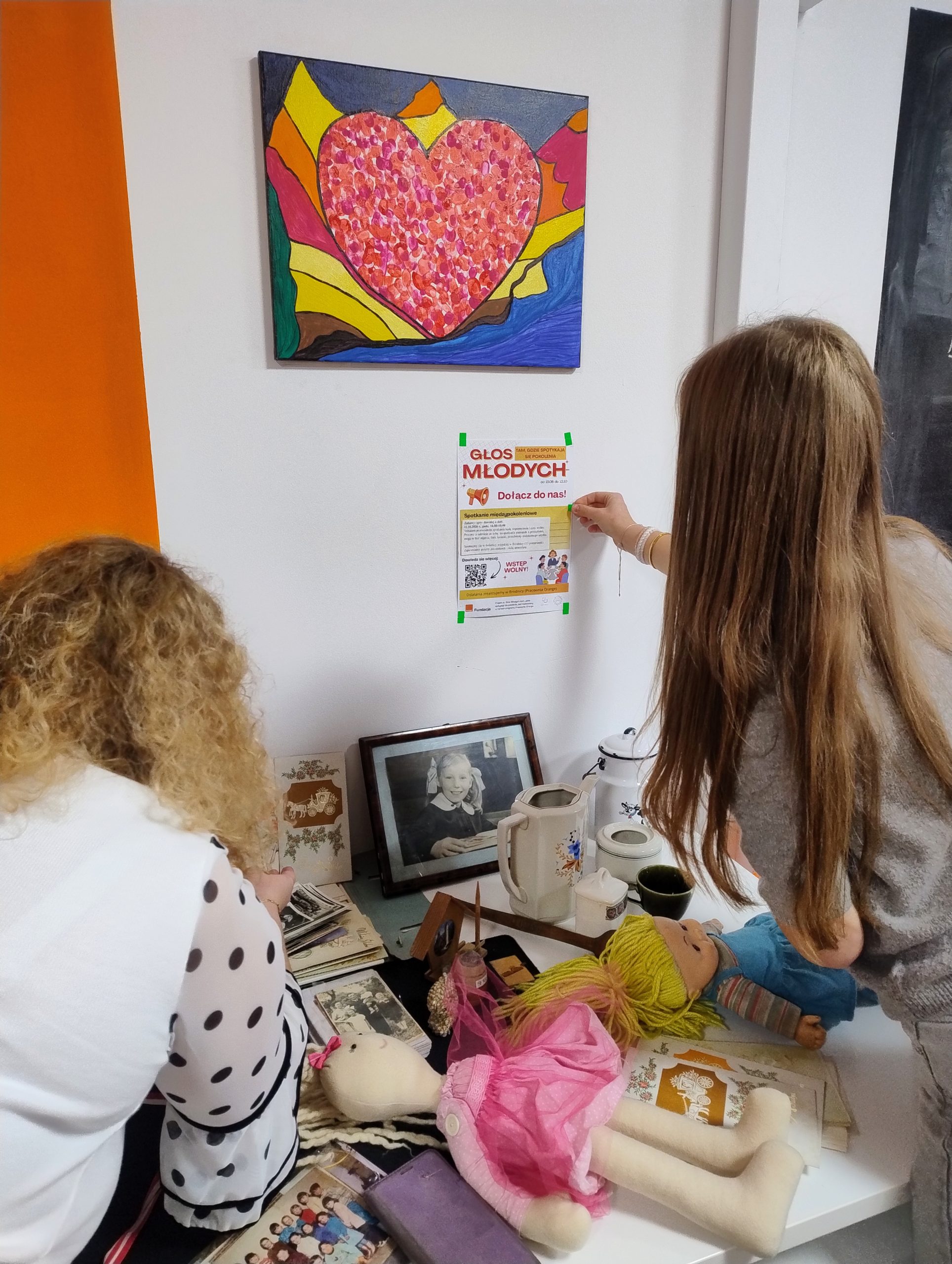 na zdjęciu liderka przykleja plakat projektu na ścianę, druga z liderek układa przedmioty z przeszłości - zdjęcia, maskotki, pamiątki