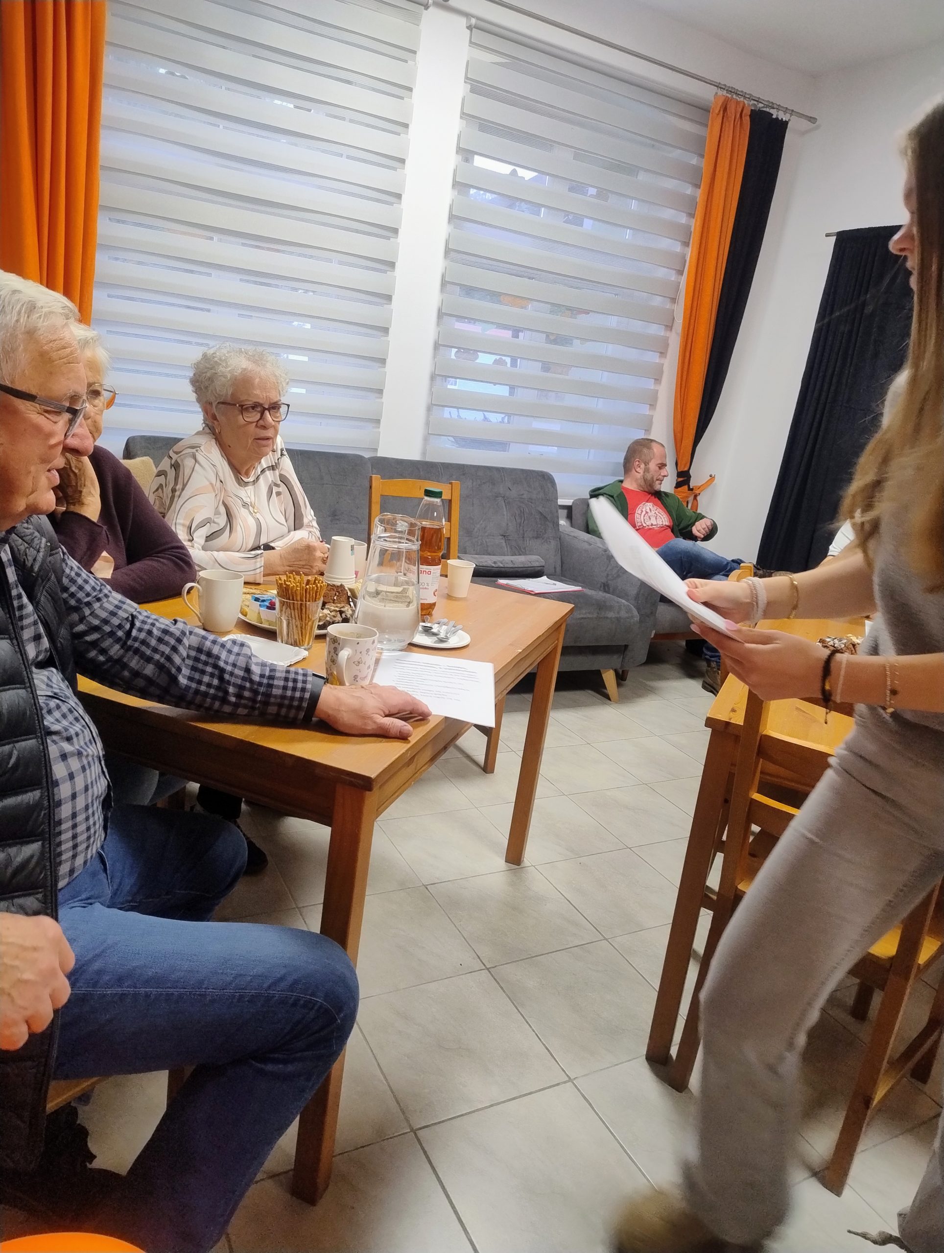 na zdjęciu widać siedzących przy stole seniorów i jedną osobę na fotelu