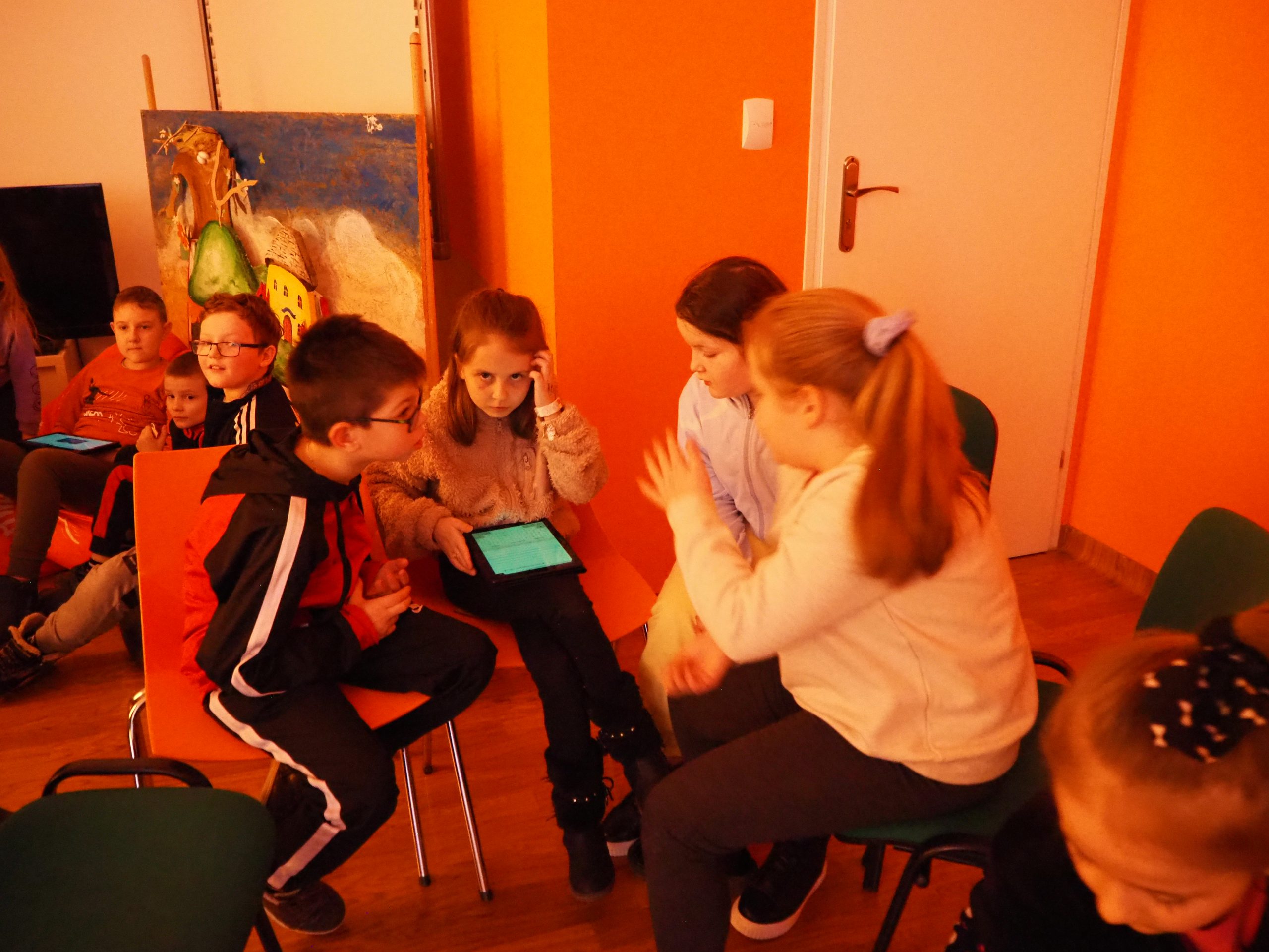 grupka dzieci w pracowni orange na tle pomarańczowej ściany korzysta z tabletu podczas zajęć edukacyjnych