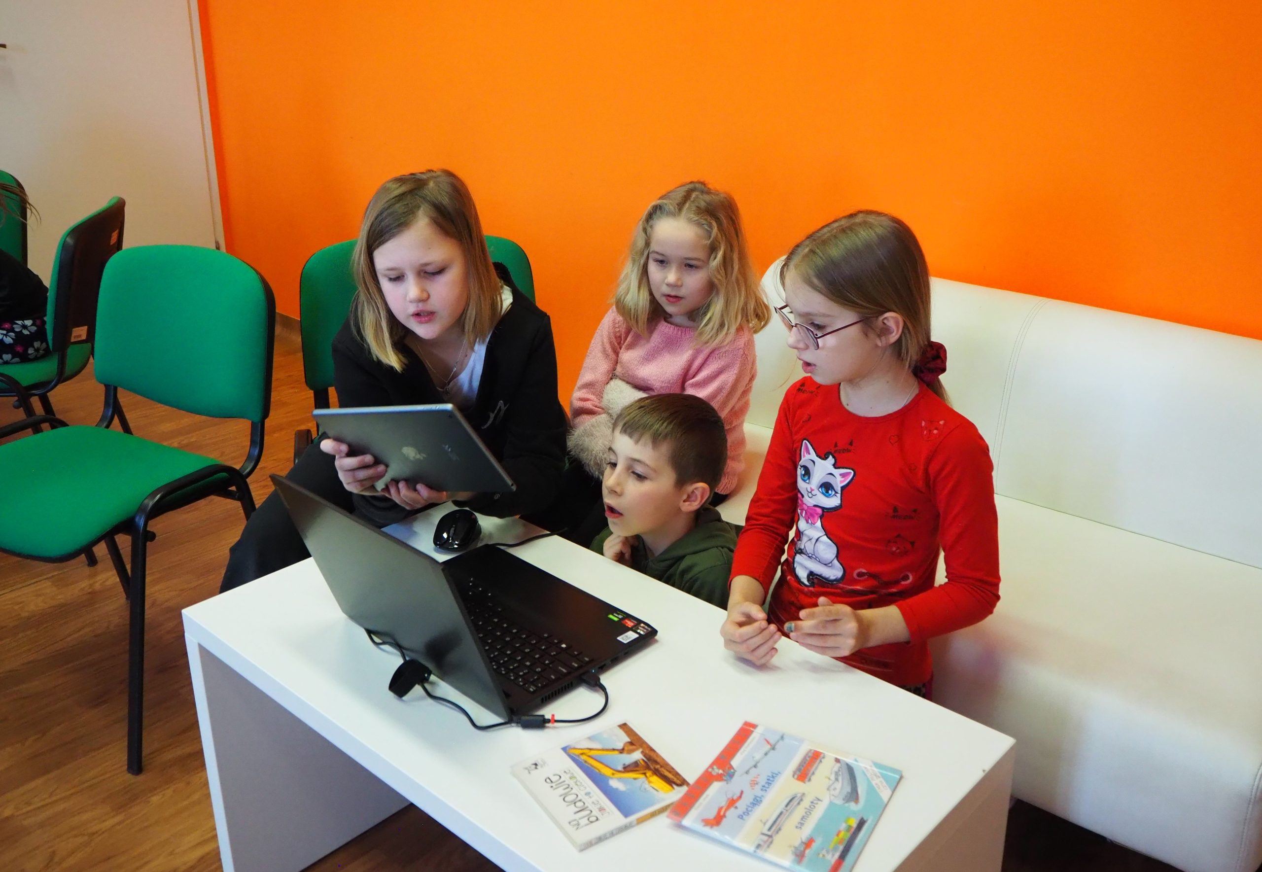 czwórka dzieci na tle pomarańczowej ściany korzyta a laptopa jedna z dziewczynek trzyma tablet