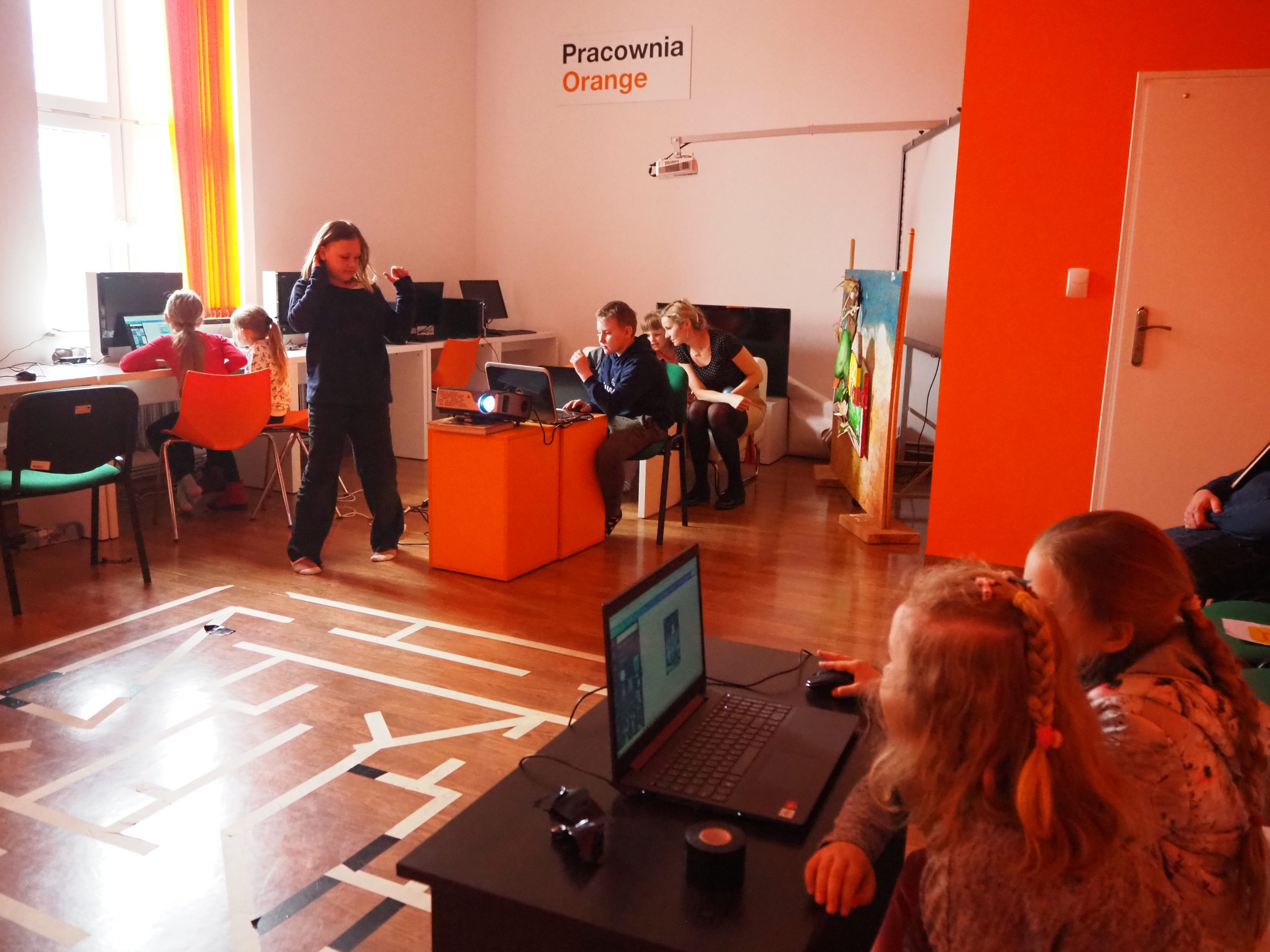 dzieci w pracowni orange siedzą przy laptopach a po środku pomieszczenia na podłodze z taśmy zrobiony jest labirynt