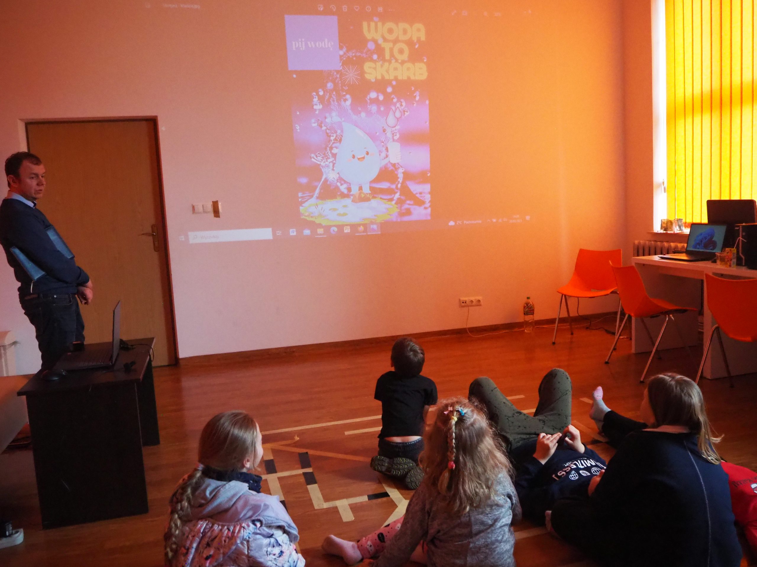 dzieci siedzą na podłodze i oglądają prezentacje wyświetlaną z projektora na ścianie, z lewej strony stoi mężczyzna