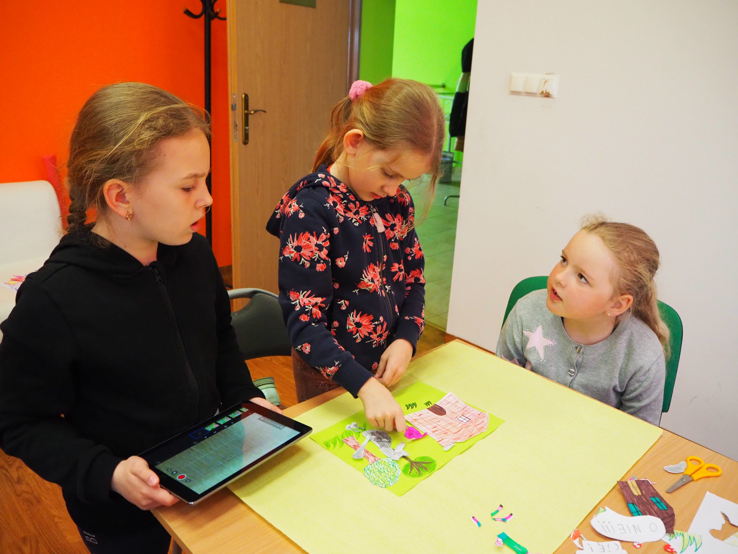 trzy dziewczynki przy stole robią razem obrazek jedna z nich trzyma tablet a druga układa elementy z papieru na zielonej kartce