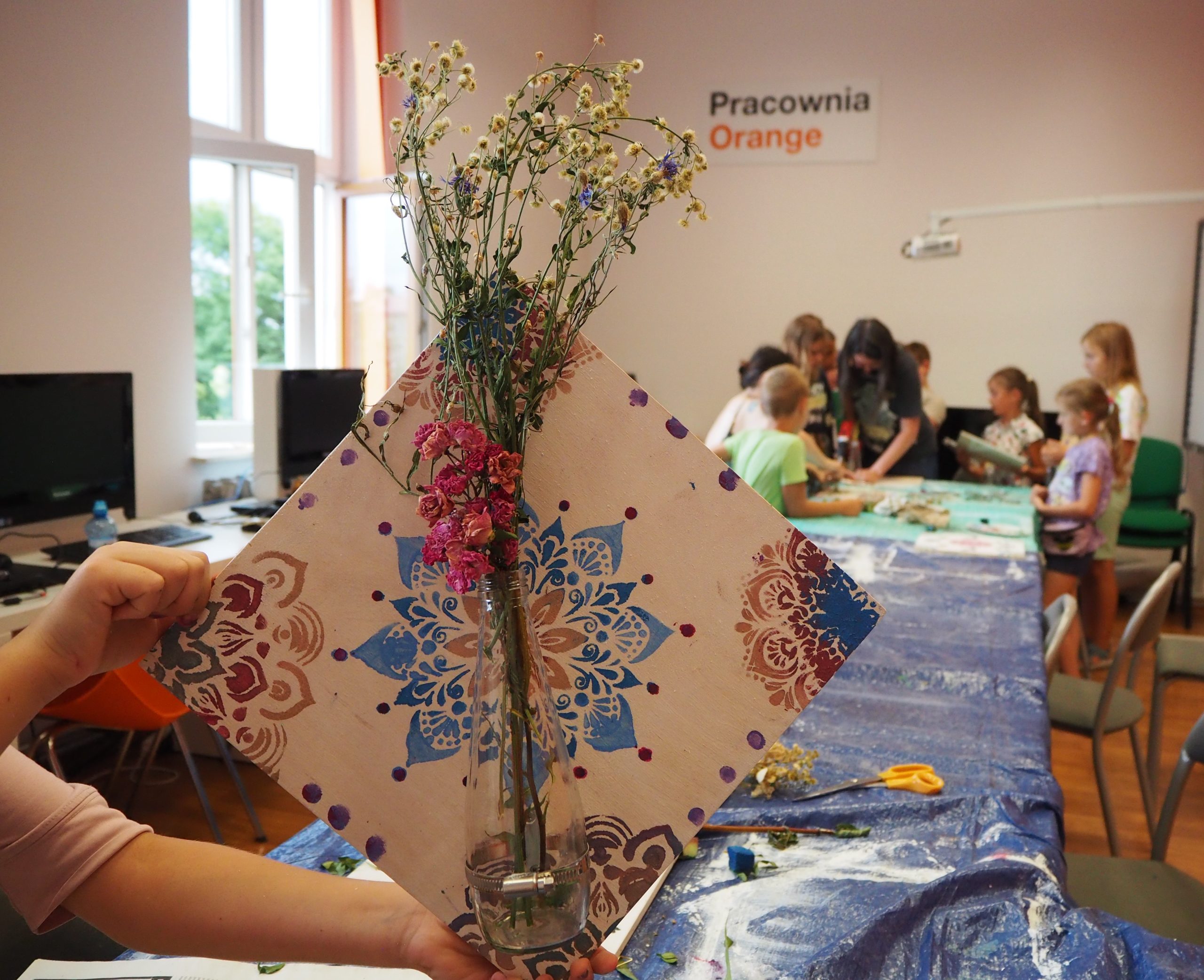 zdjęcie przedstawia pracę jednej z uczestniczek, pomalowana na różne wzory płytka z drewna a na nim doczepiony wazonik z kwiatami