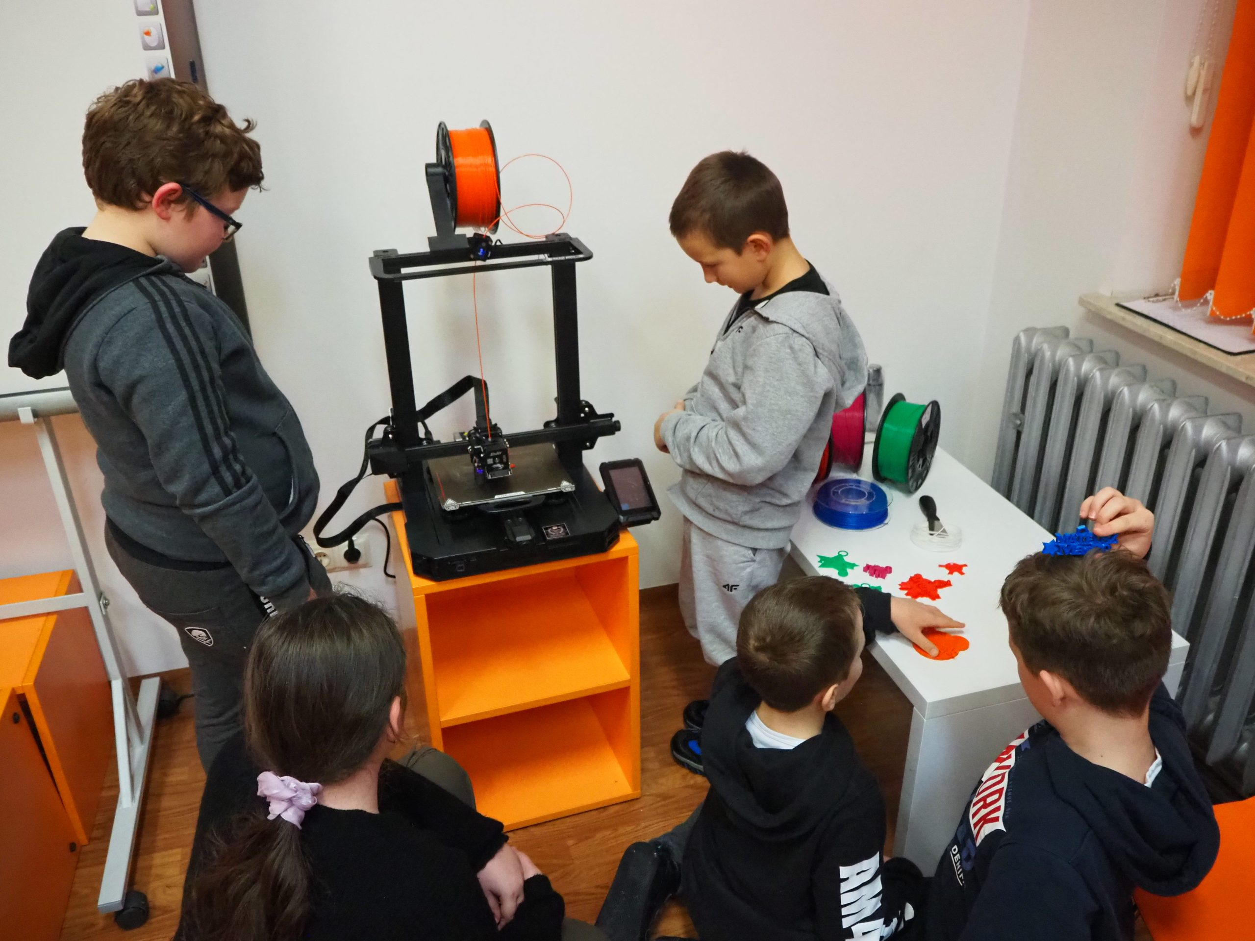 Grupa dzieci obserwuje proces drukowania na drukarce 3D.