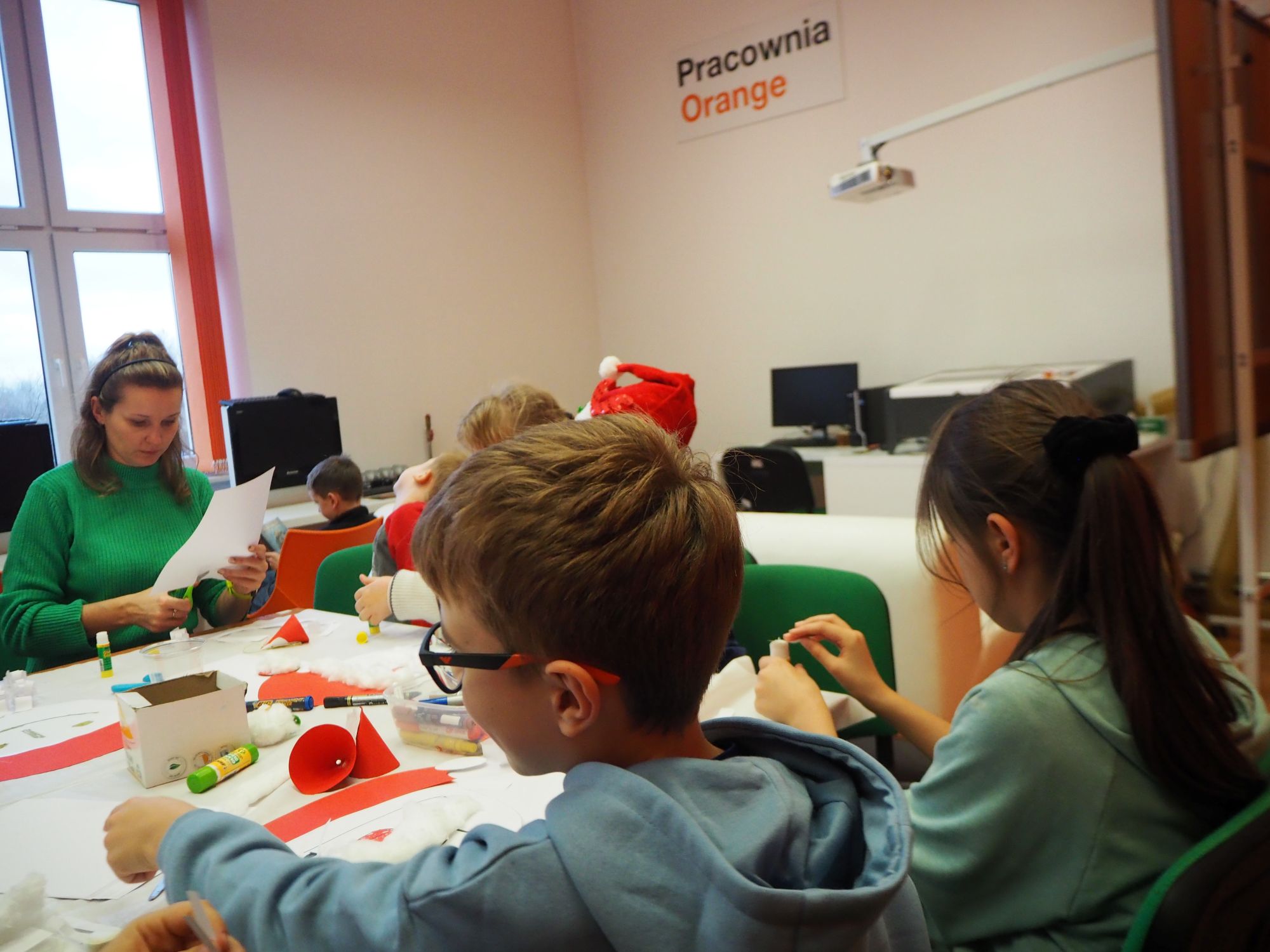 dzieci siedza przy stole i z Panią prowadzącą zajęcia wykonują mikołaje z papieru