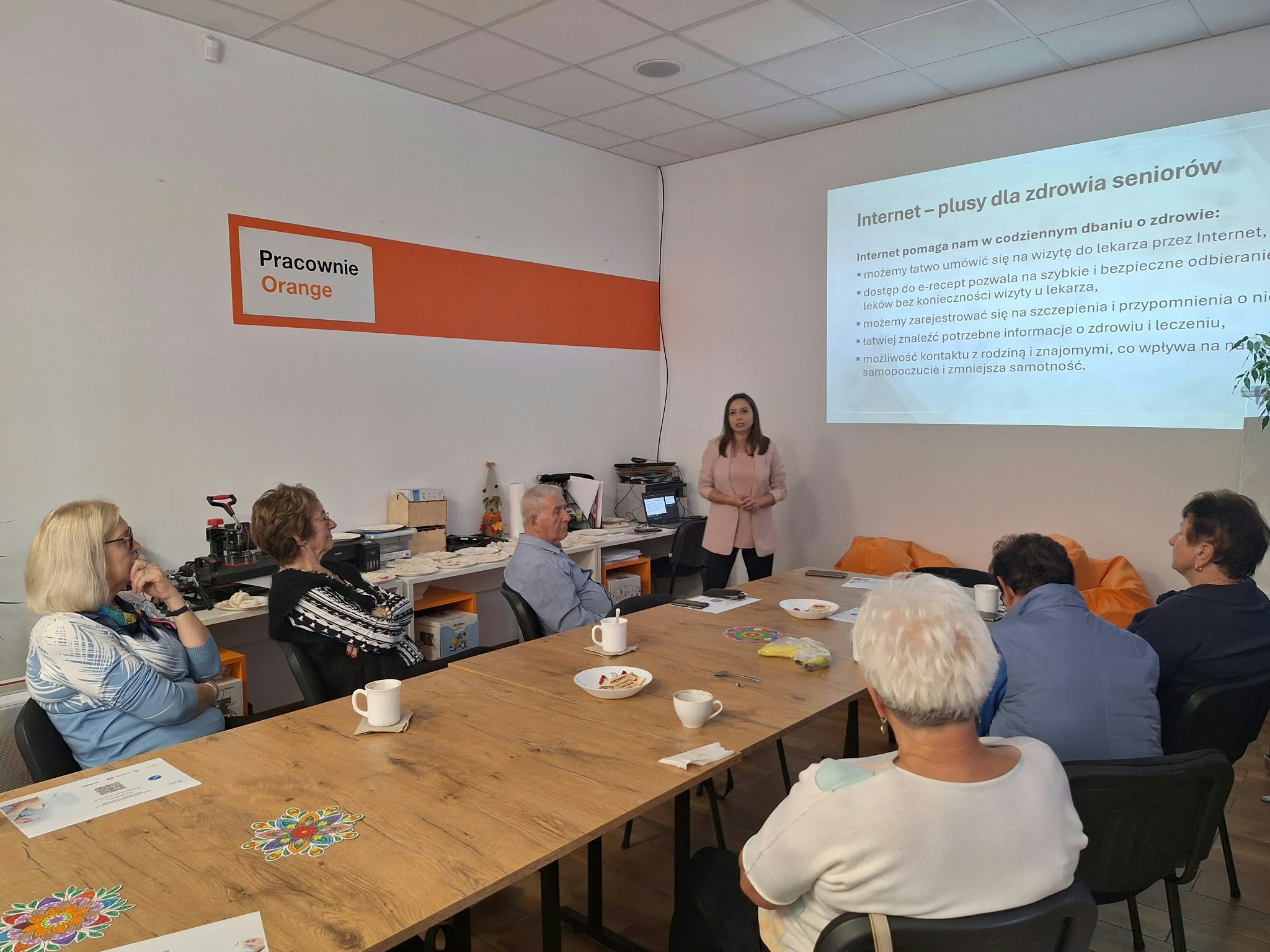 Grupa osób w Pracowni Orange - seniorzy słuchają wykładu.
