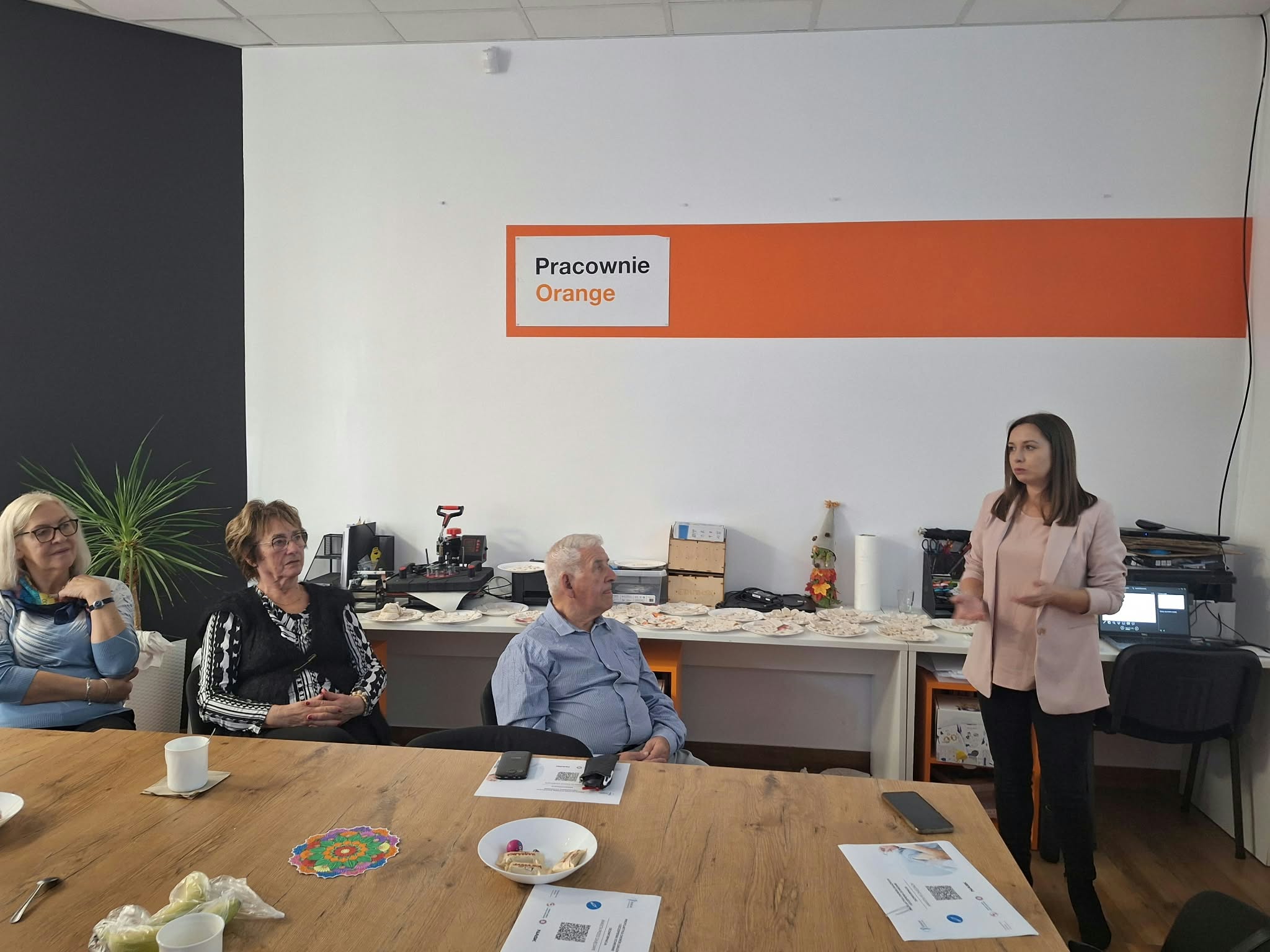 Grupa osób w Pracowni Orange - seniorzy słuchają wykładu.