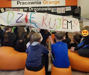 Pomieszczenie, w którym dzieci i młodzież siedzący tyłem na pomarańczowych pufach i trzymający transparent z napisem Dziękujemy! Siedzą na tle ściany z widoczną na jej górze nazwą Pracownia Orange w Drzonowie
