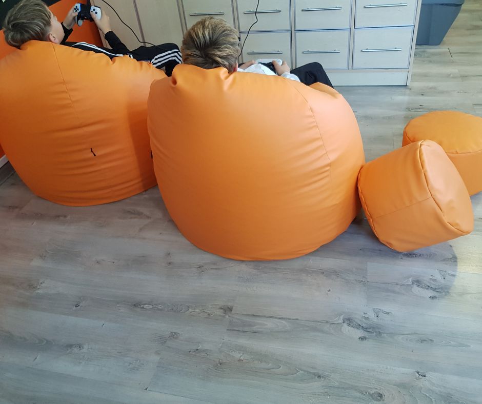 Pomieszczenie, na pierwszym tle widoczny tył dwóch młodych osób, siedzących na pomarańczowych, dużych pufach przed dużą komodą z szufladami. Obok nich, z prawej strony dwie małe pufy. Osoby mają w dłoniach kontrolery do gry.