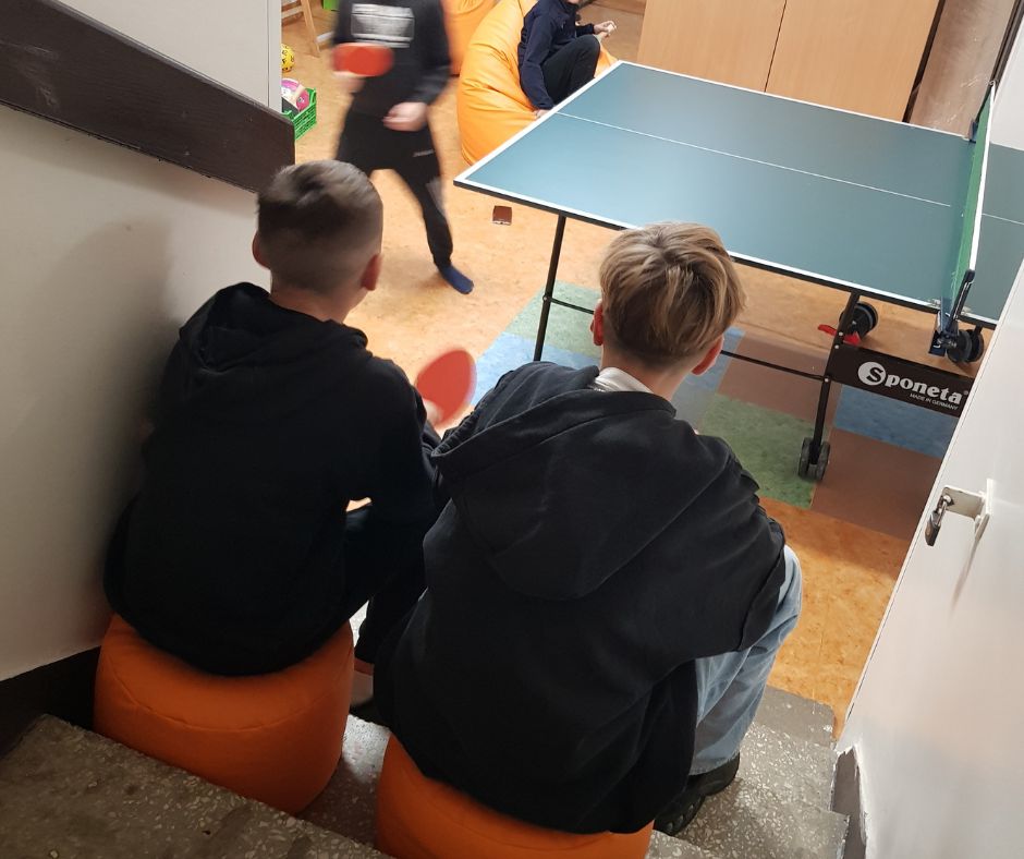 Pomieszczenie, a w nim dwie młode oby siedzące tyłem na małych pomarańczowych pufach, patrzące na osoby grające w ping ponga przy stole.