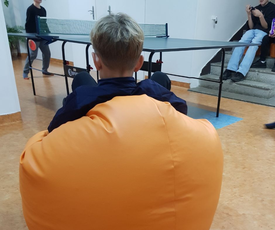 Sala, a w niej na pierwszym planie młoda osobą siedząca tyłem na dużej, pomarańczowej pufie. W tle stół do ping ponga z grającym.