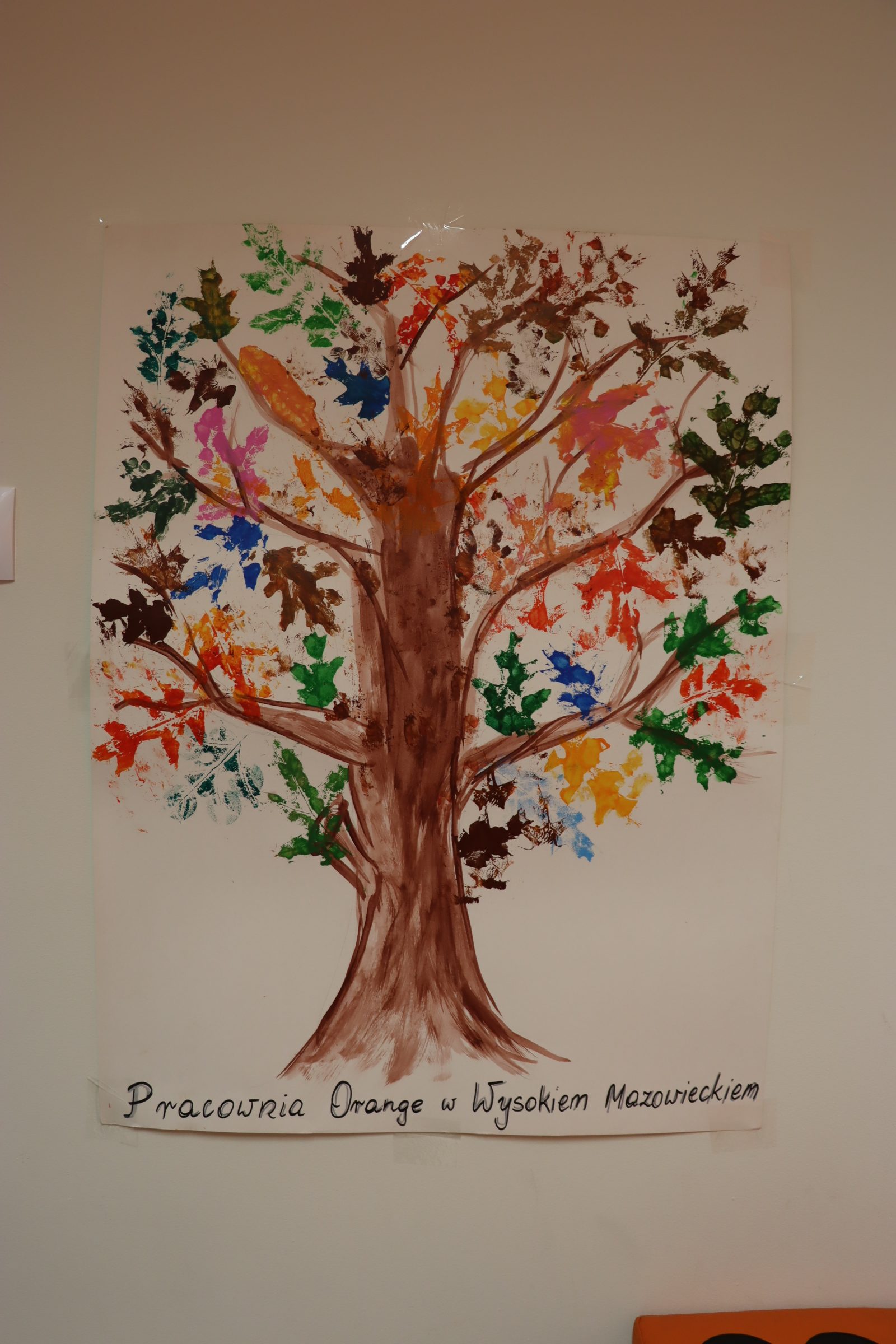 Zdjęcie pracy plastycznej, którą stworzyły dzieci wraz z animatorką. Jest to drzewo z kolorowymi liśćmi. Pod spodem znajduje się napis Pracownia Orange w Wysokim Mazowieckiem.