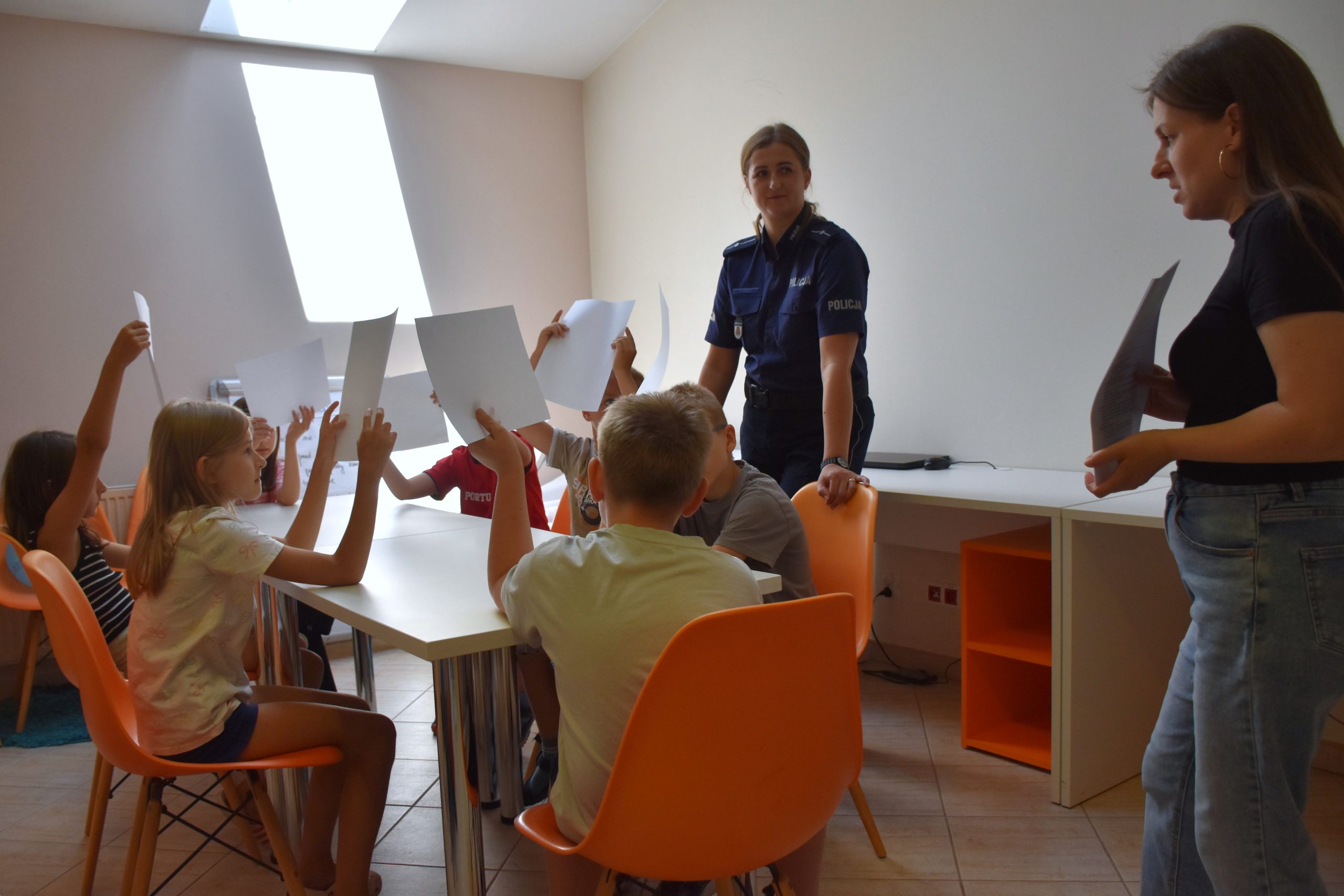 dzieci siedzą przy stole i podnoszą kartki, przed nimi stoi policjantka