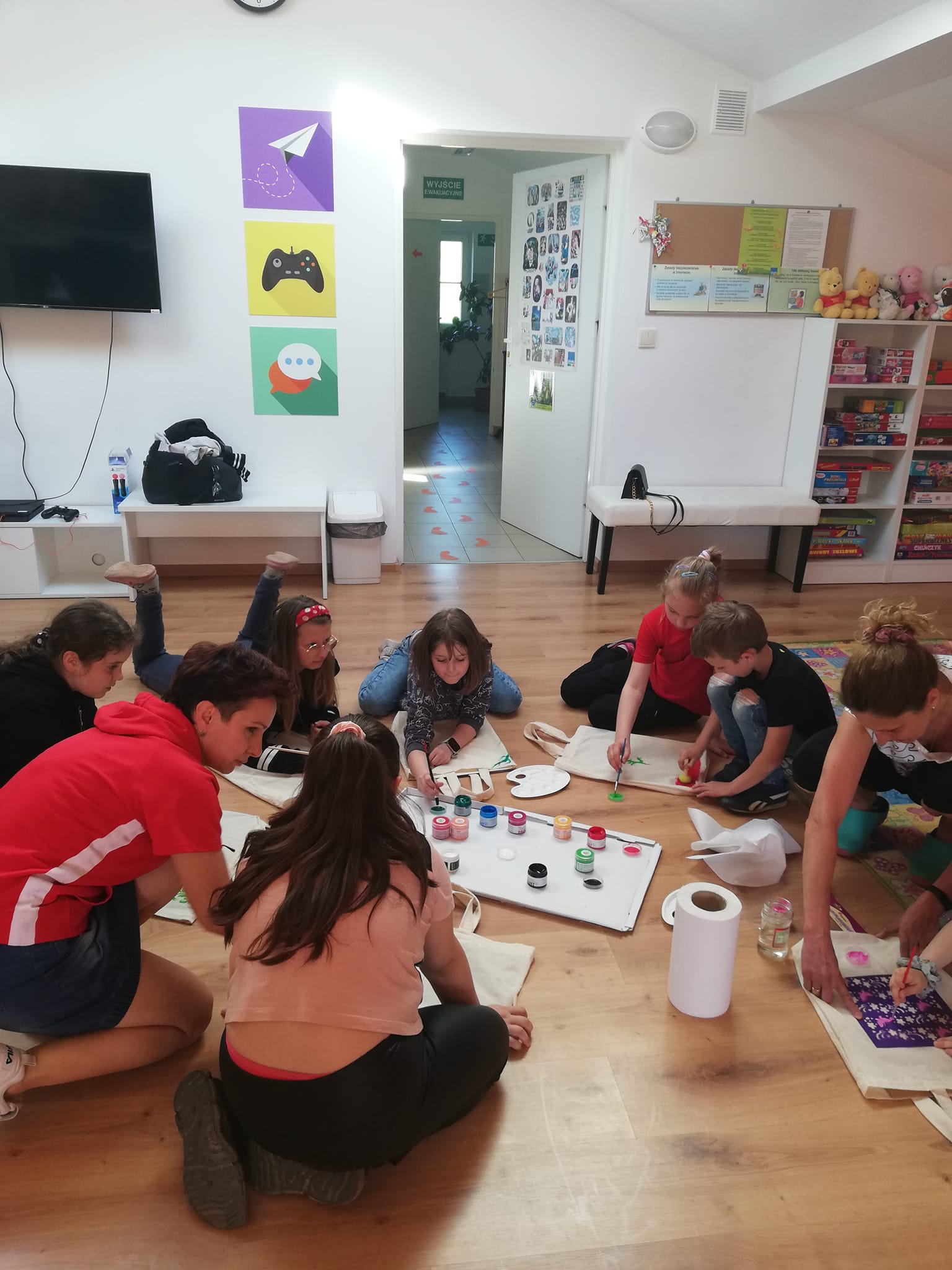 Grupa dzieci i młodzieży siedzących na podłodze, malują farbami na torbach bawełnianych.