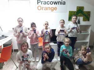 Na zdjęciu jest dziewięcioro dzieci, trzymają w rękach kolorowe sakiewki. Nad ich głowami jest napis Pracownia Orange.