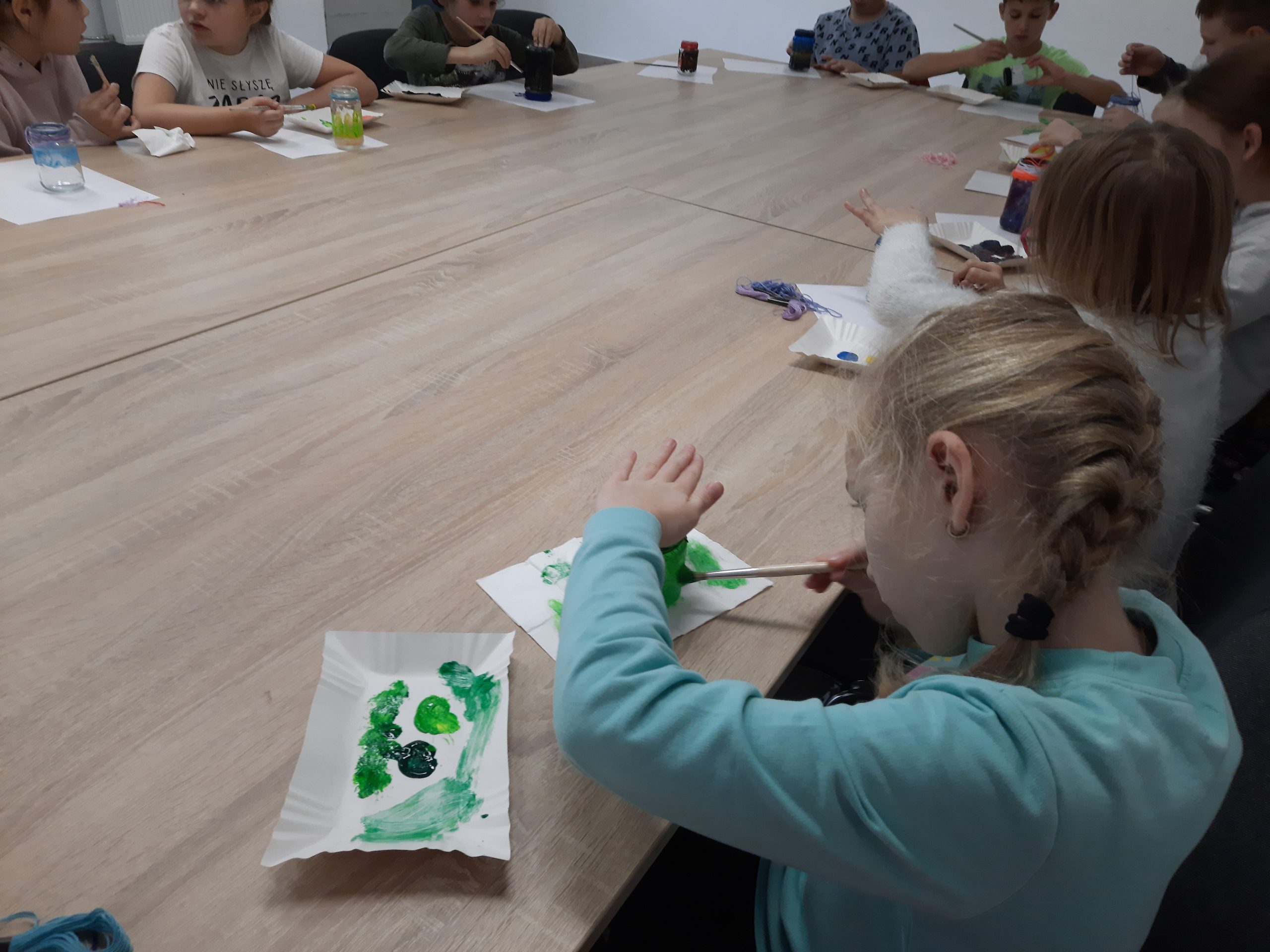 Na obrazku widoczne są dzieci owijające słoiki kolorowymi sznurkami. Na pierwszym planie dziewczynka w warkoczu owija słoik zieloną nitką.