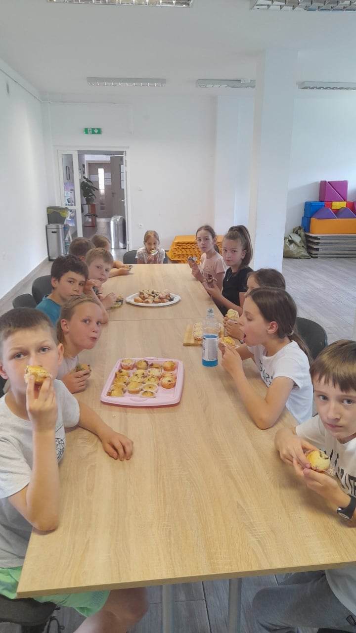 Na zdjęciu widoczna jest grupa dzieci siedząca przy stole i jedząca muffinki.